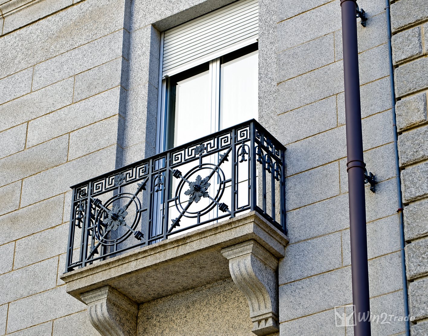 Un balcon en granit orné d'un garde-corps en fer forgé à motifs de grecques et de rosaces, sous une fenêtre à volet roulant.