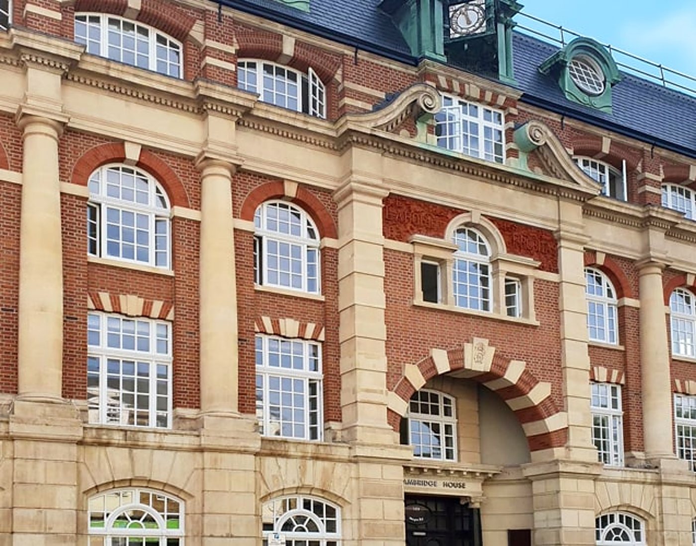 Façade d'un grand bâtiment ancien en briques rouges et pierre beige, avec de hautes fenêtres cintrées, une entrée en arc et un toit en ardoise.