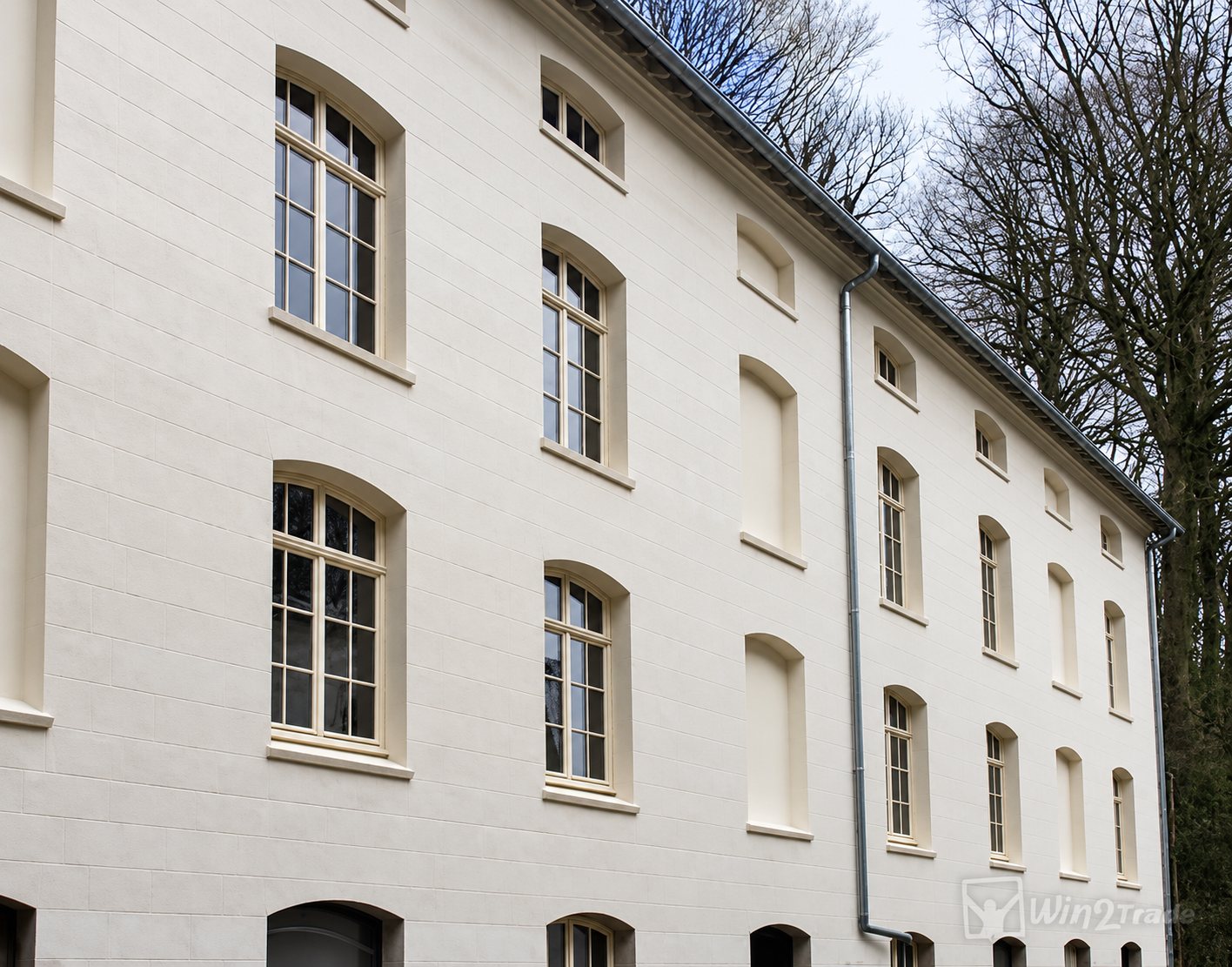 Façade d'un bâtiment ancien rénové, enduite de blanc, avec de hautes fenêtres cintrées à petits carreaux et des arbres nus en arrière-plan.