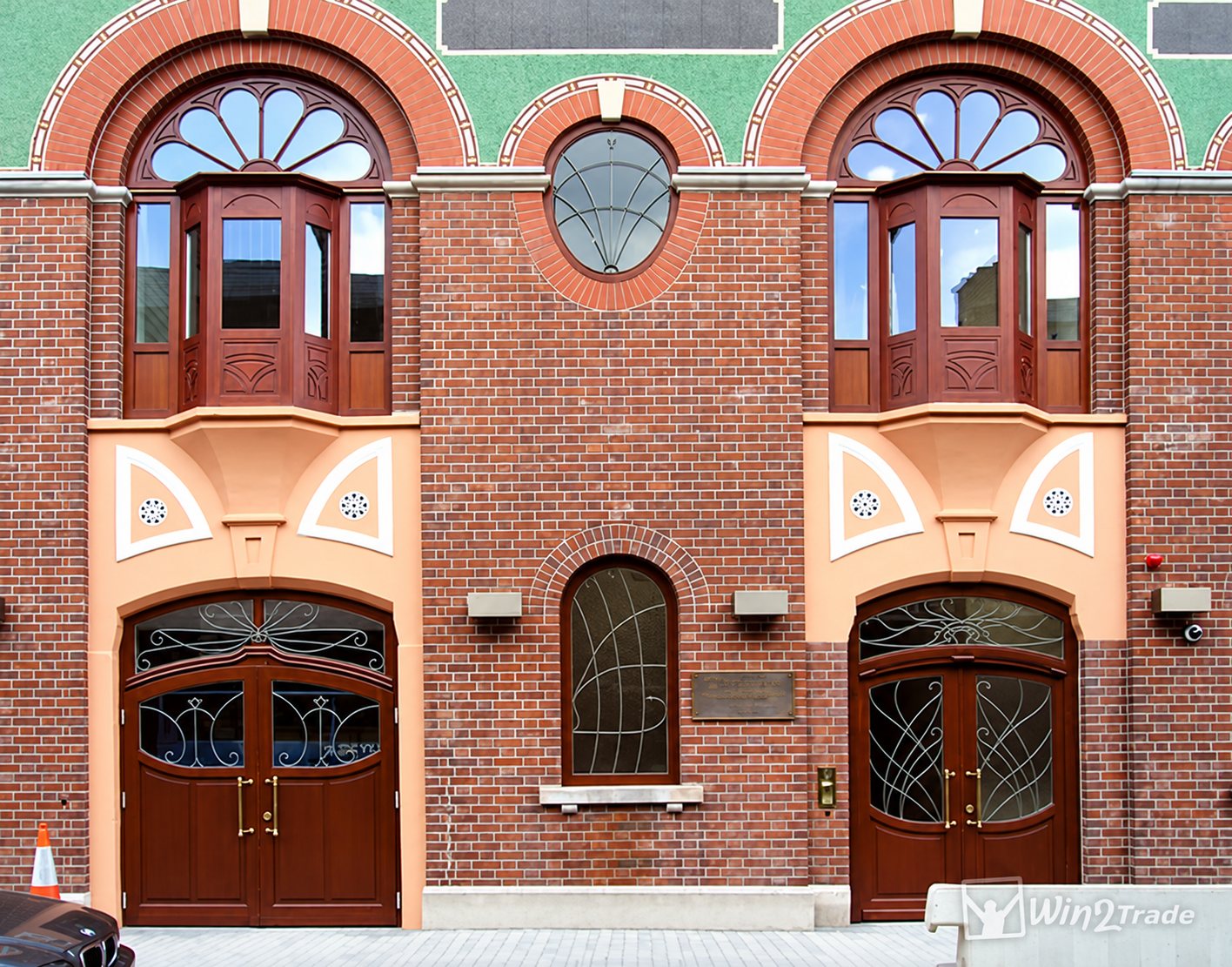 Façade d'un bâtiment en briques rouges avec deux oriels en bois, des portes cintrées en bois sombre et des décorations orange et vertes.