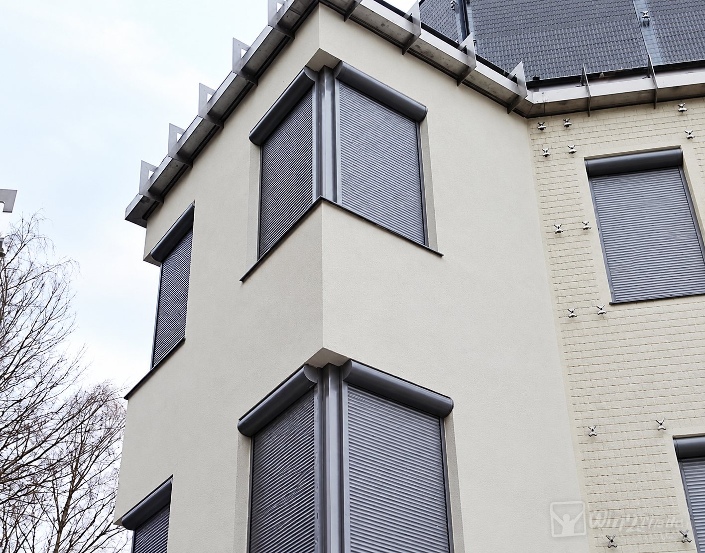 Façade claire d'un immeuble dont les fenêtres d'angle sont équipées de volets roulants gris fermés.