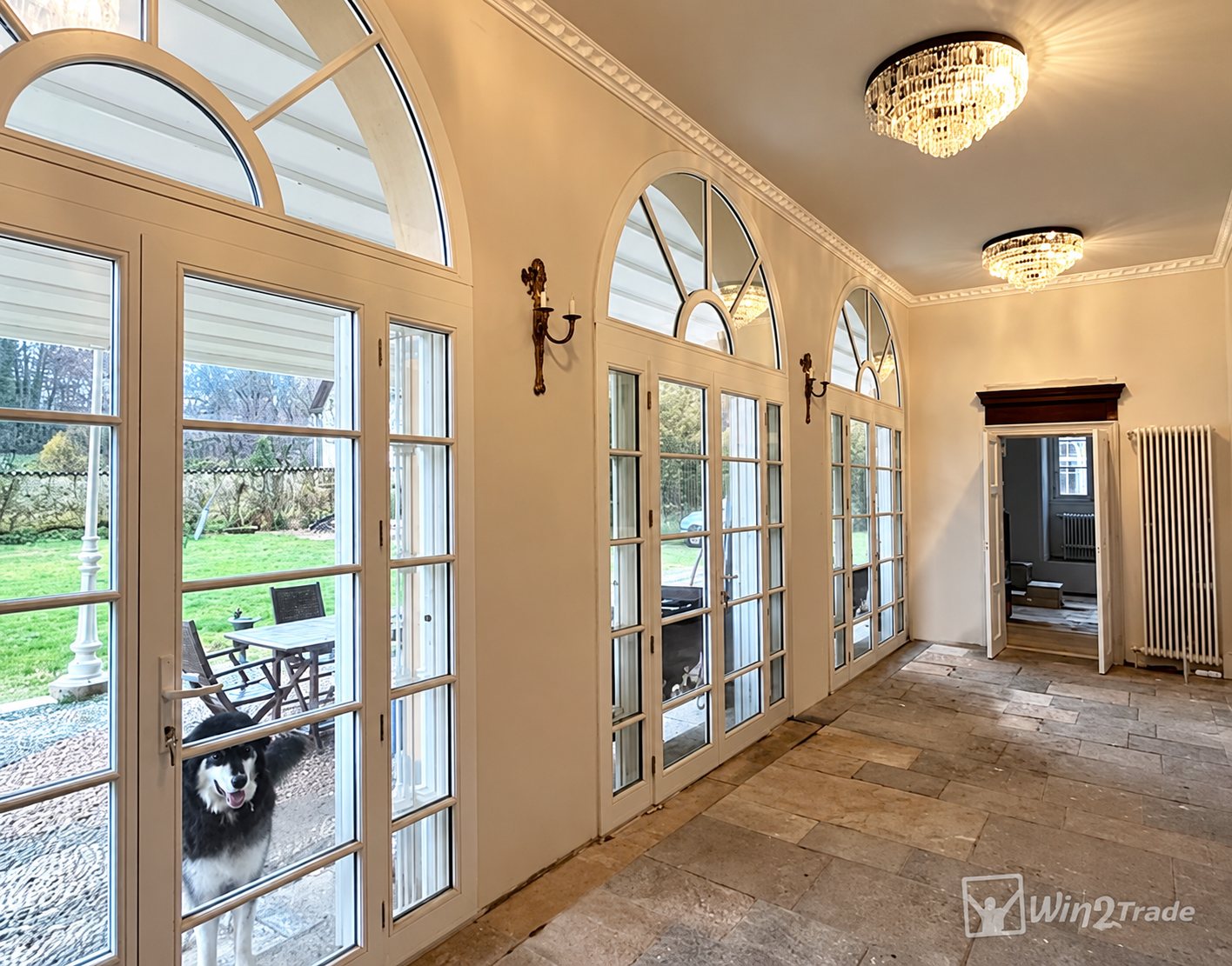 Une galerie intérieure au sol en dalles de pierre, dotée de grandes portes-fenêtres cintrées blanches donnant sur un jardin, avec des lustres en cristal et un chien qui regarde à travers la vitre.