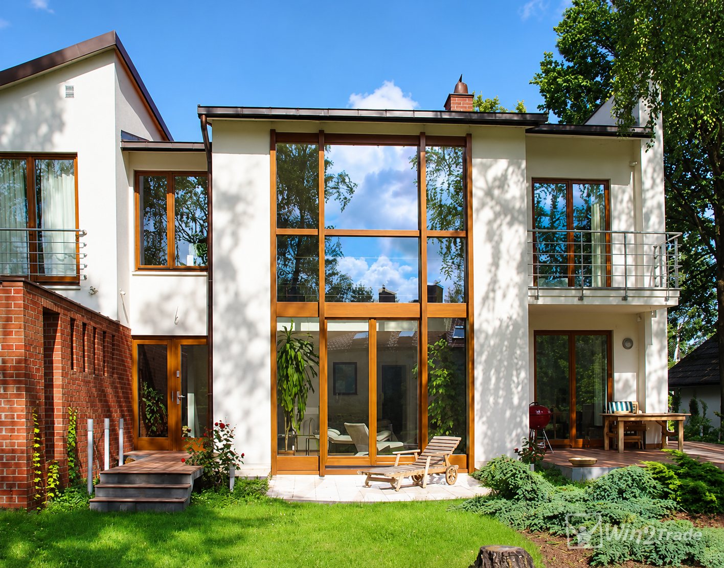 Façade arrière d'une maison blanche moderne avec une grande baie vitrée à cadre en bois, un balcon, une terrasse et un jardin verdoyant.