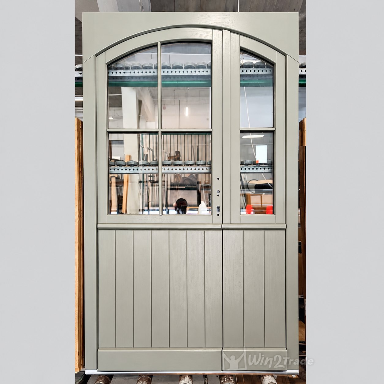 Une porte double en bois peint gris clair, au sommet cintré, avec une partie haute vitrée à petits carreaux et un bas à lames verticales, photographiée dans un atelier.