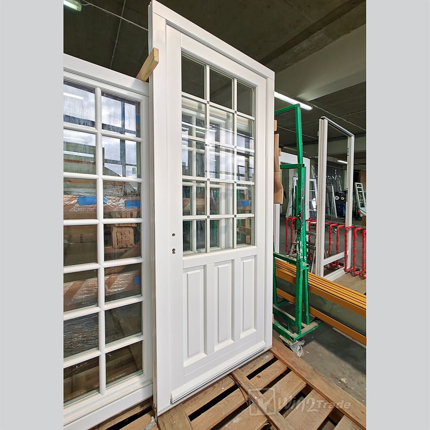 Une porte d'entrée en bois blanche, avec vitrage à petits carreaux et panneaux en bas, posée sur une palette dans un atelier, à côté d'une fenêtre blanche.