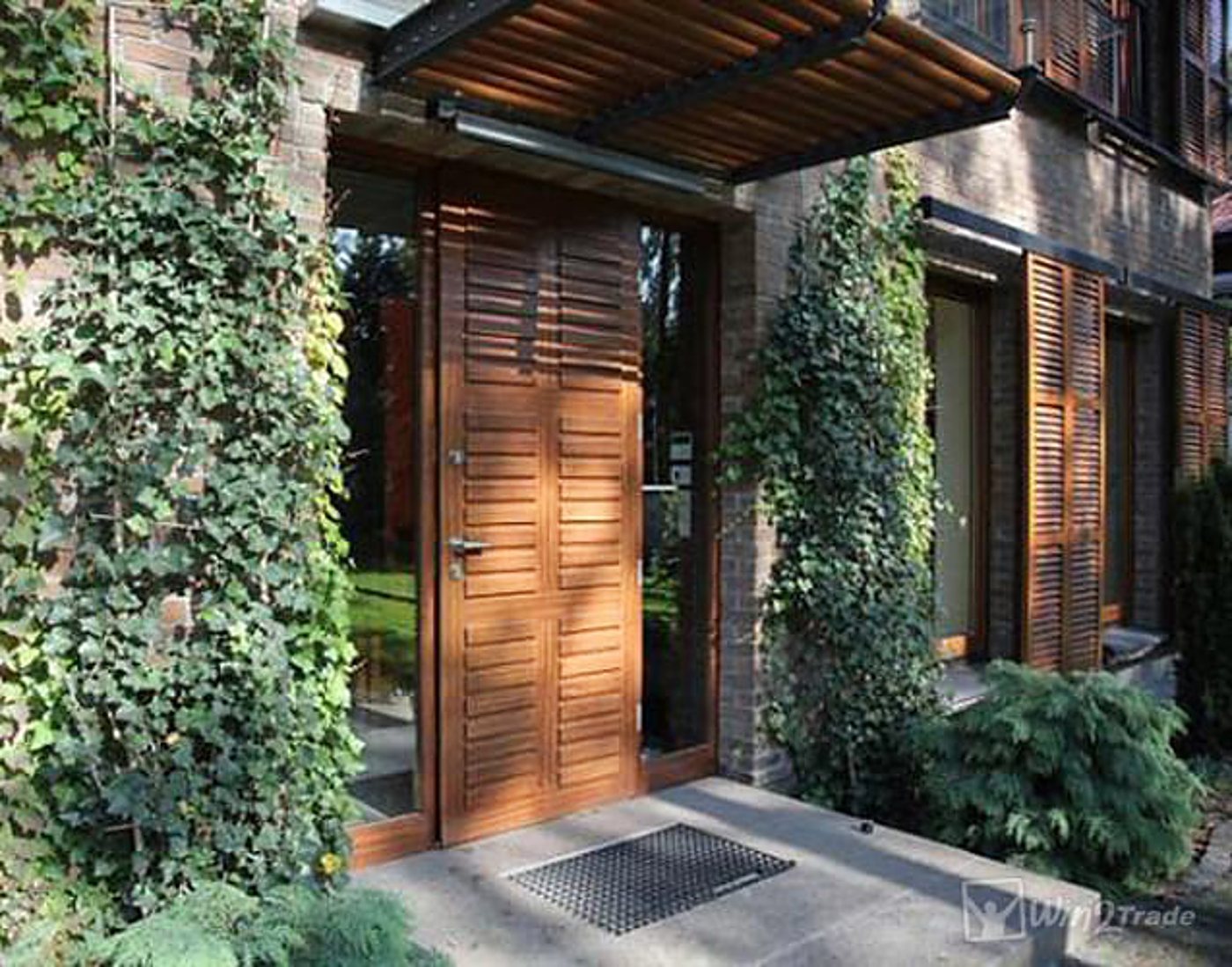 Une porte d'entrée en bois à lattes horizontales, encadrée de vitrages, sous un auvent en bois, sur une façade en brique couverte de lierre.