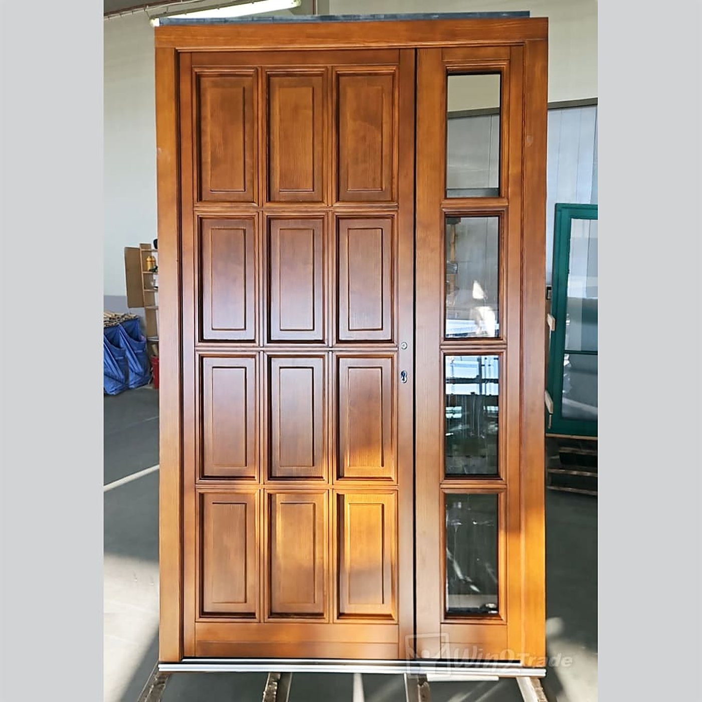 Porte d'entrée en bois teinté miel composée de douze panneaux et d'un côté vitré, exposée dans un atelier.