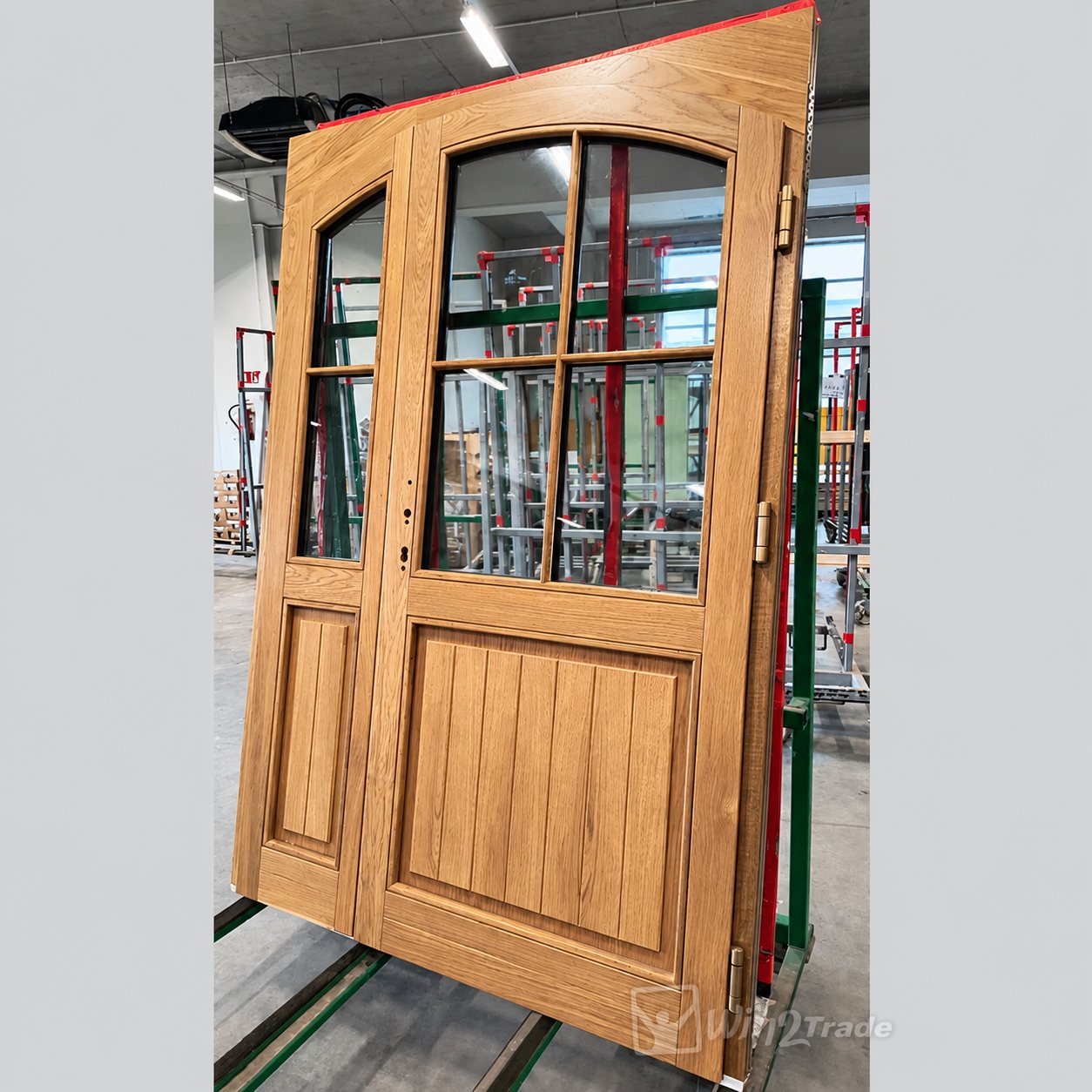 Une porte d'entrée à deux vantaux en chêne clair, avec petits-bois et partie haute vitrée cintrée, posée sur un chariot dans un atelier de fabrication.