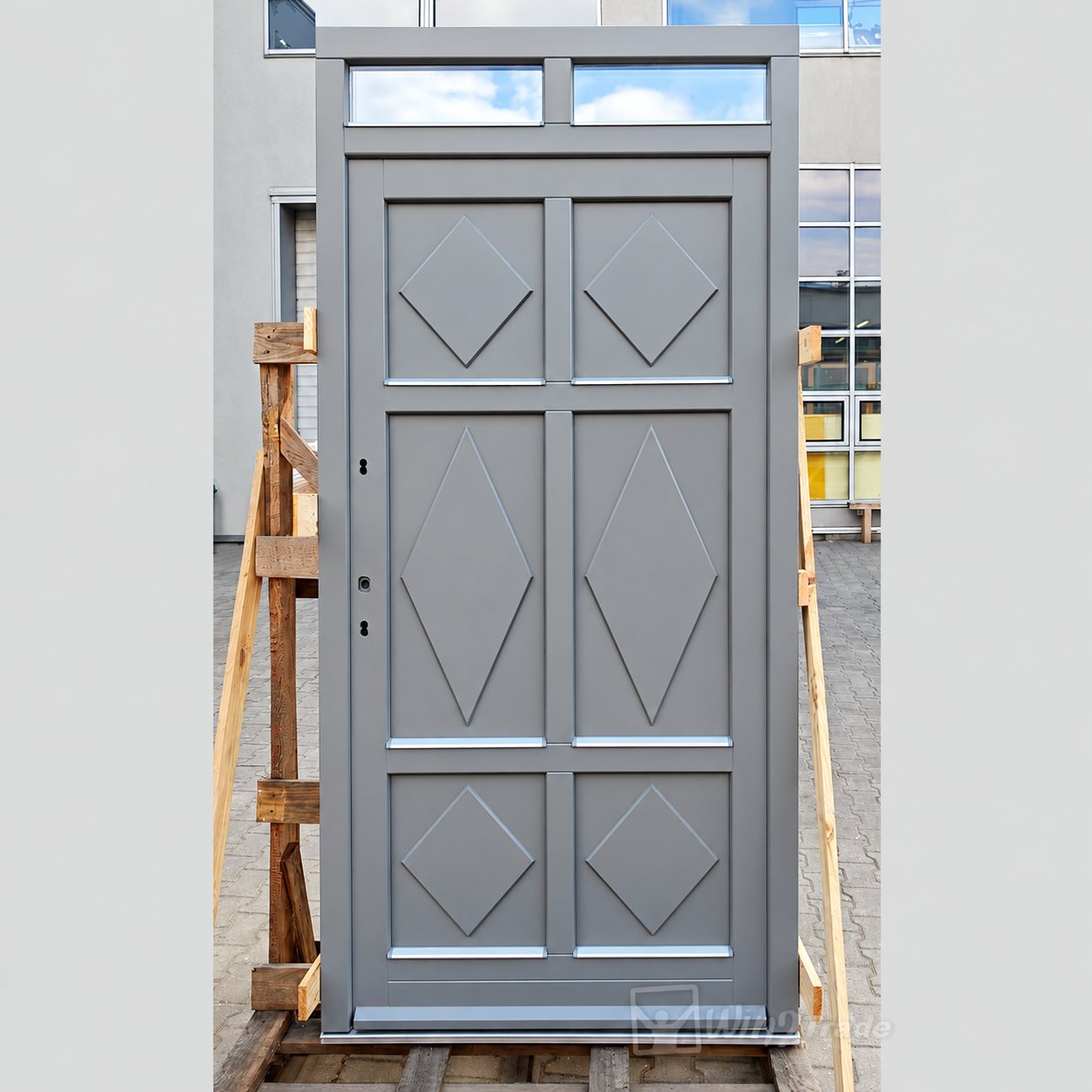 Porte d'entrée en bois peinte en gris avec panneaux à motifs de losanges et imposte vitrée, maintenue par des cales en bois devant un bâtiment.
