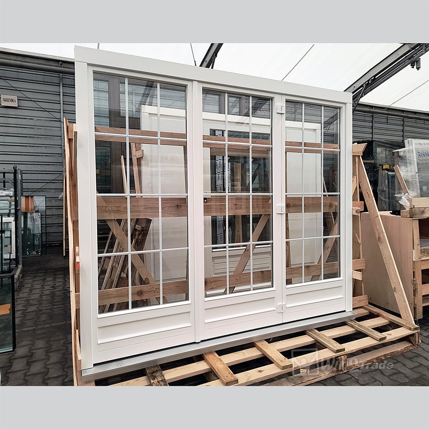 Une grande porte-fenêtre blanche à trois vantaux et petits carreaux, posée sur une palette en bois dans un entrepôt couvert.