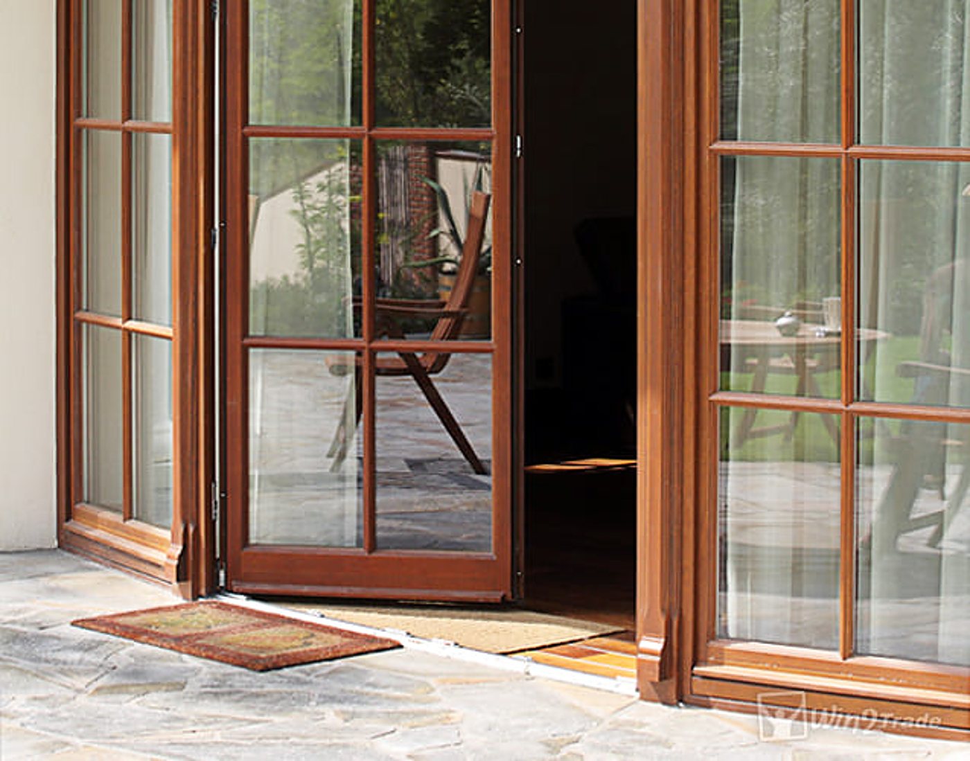 Une porte-fenêtre en bois à petits carreaux, entrouverte, donne sur une terrasse dallée de pierre avec un paillasson et des chaises de jardin.