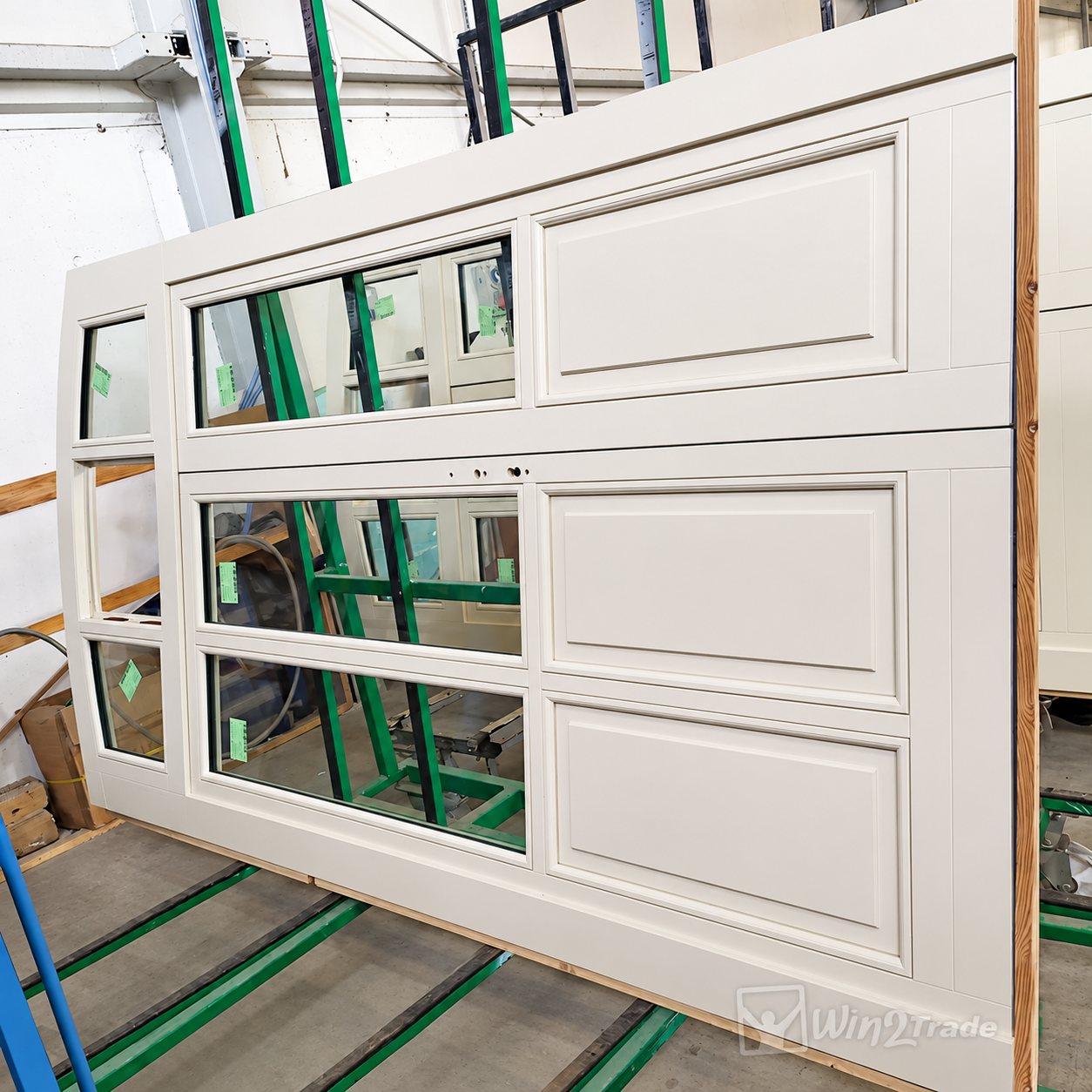 Une grande porte de garage en bois peint en blanc, composée de panneaux moulurés et de vitrages, posée sur un chariot vert dans un atelier de fabrication.