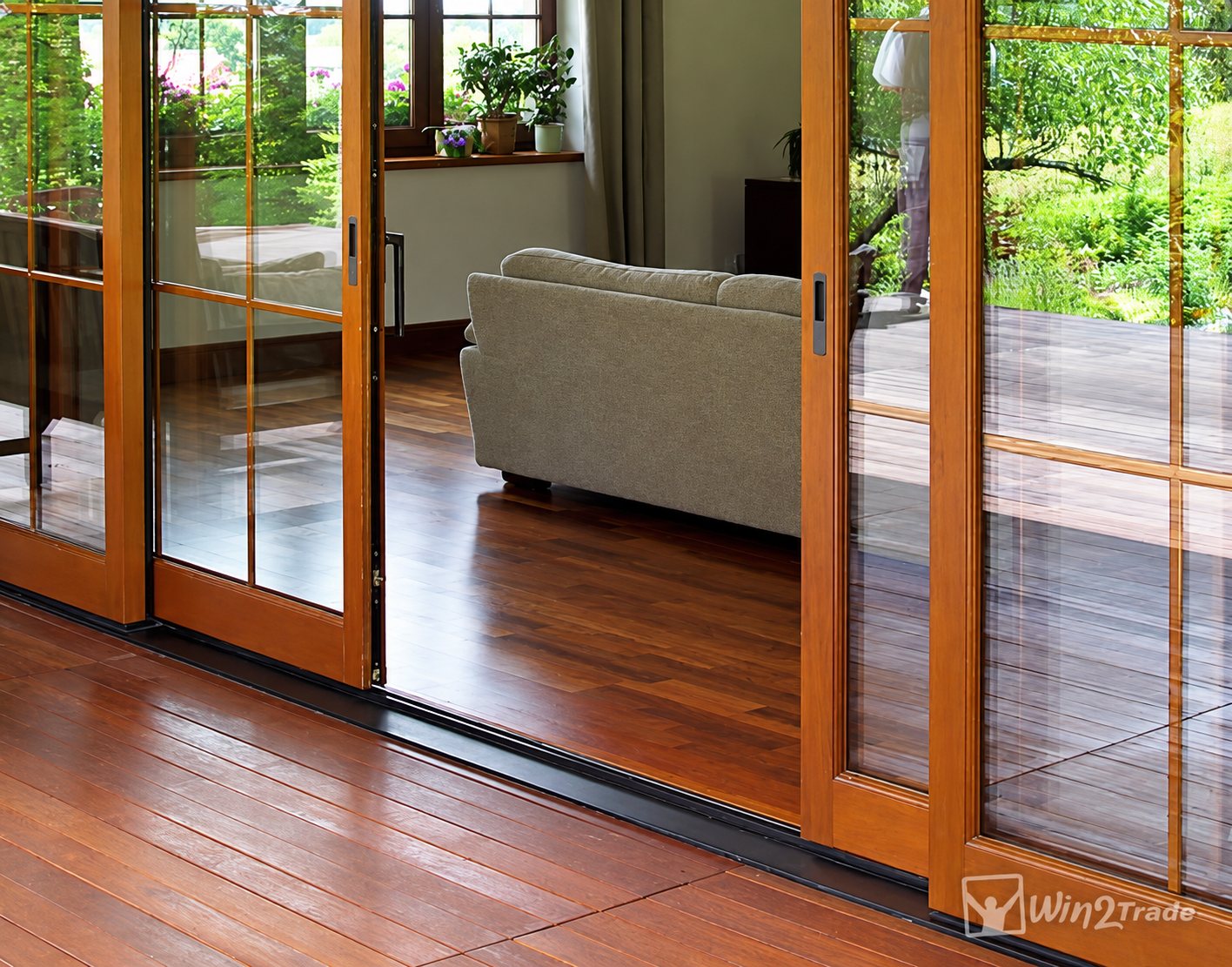 Des portes coulissantes vitrées à cadre en bois ouvrent sur un salon avec canapé gris-vert et parquet foncé, prolongé par une terrasse en bois.