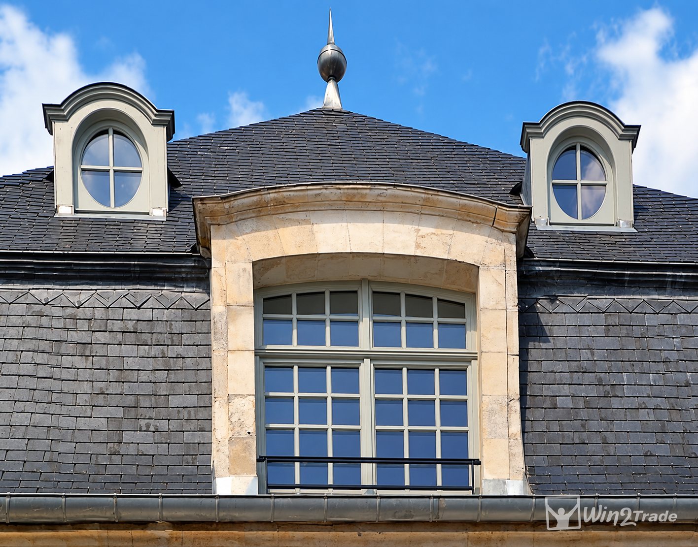 Façade de bâtiment ancien avec toit en ardoise, trois lucarnes aux encadrements de pierre claire et un épi de faîtage métallique sous un ciel bleu.