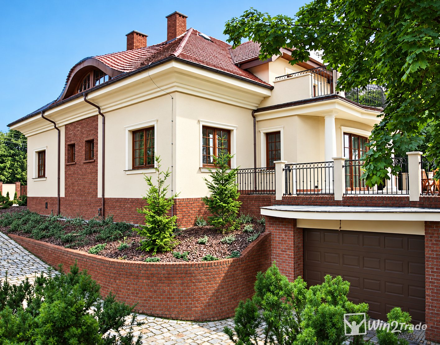 Une grande villa claire au toit de tuiles rouges, avec soubassement en brique, terrasse à balustrade, porte de garage brune et jardin arbustif au premier plan.